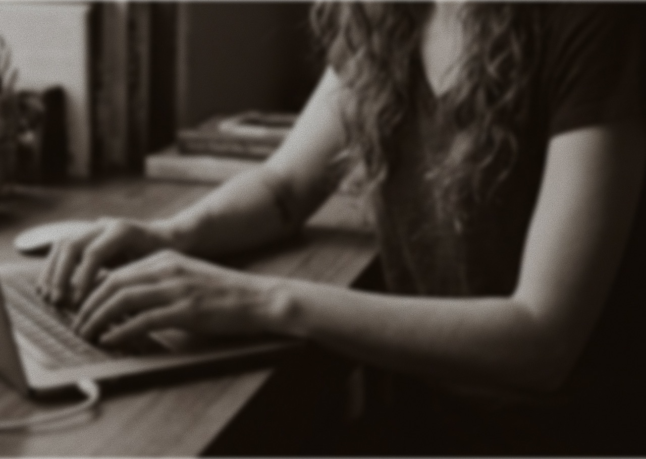 Black and white image of Jessica, founder of Partial History, sitting at a desk and typing on a laptop. The image is cropped to show only her arms and torso.
