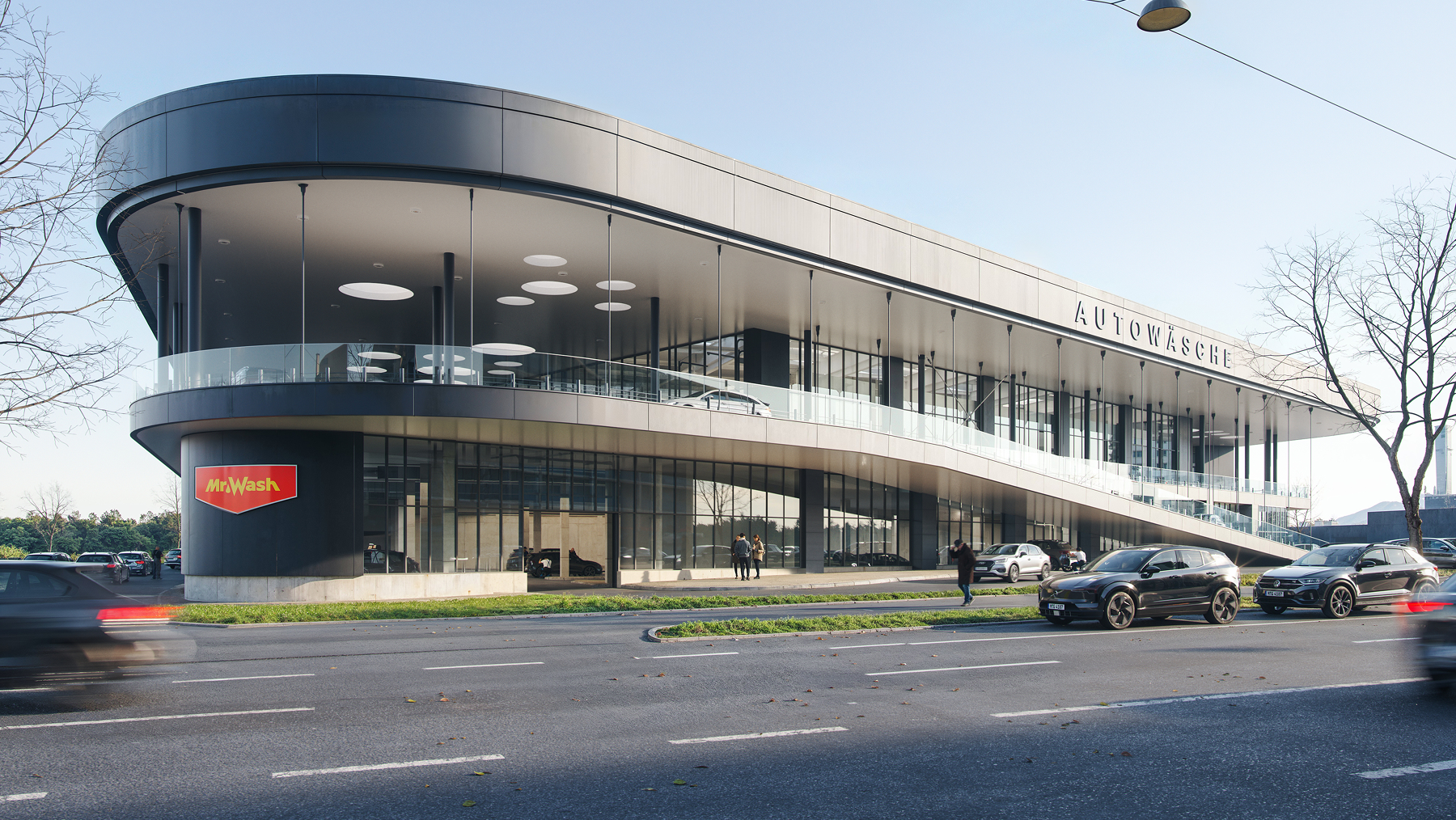 Modern, two-story car wash building with curved design, glass walls, and ramp with cars near a multi-lane street.
