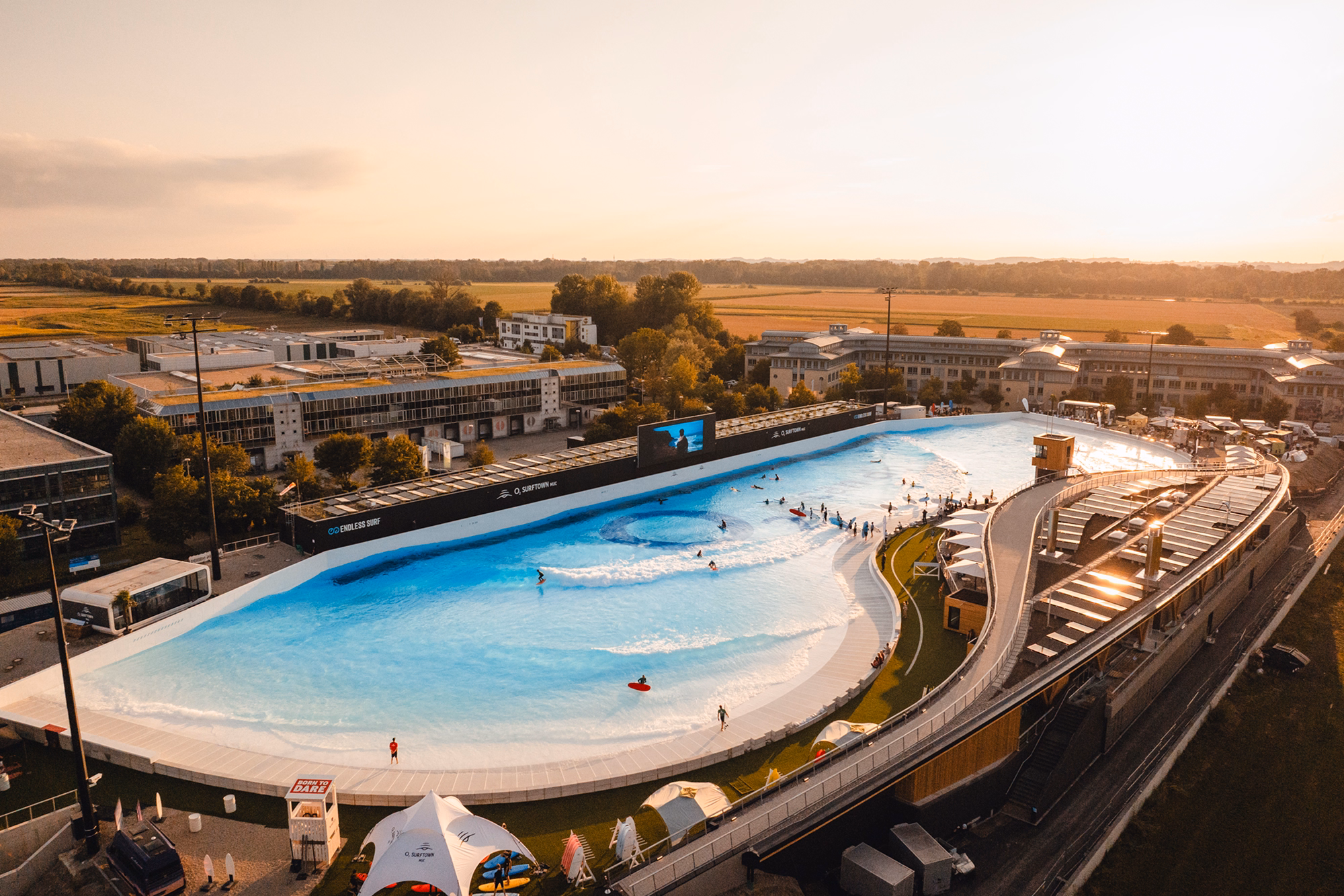 Aerial view of an outdoor artificial surfing pool with people surfing and watching, surrounded by buildings and green fields at sunset.