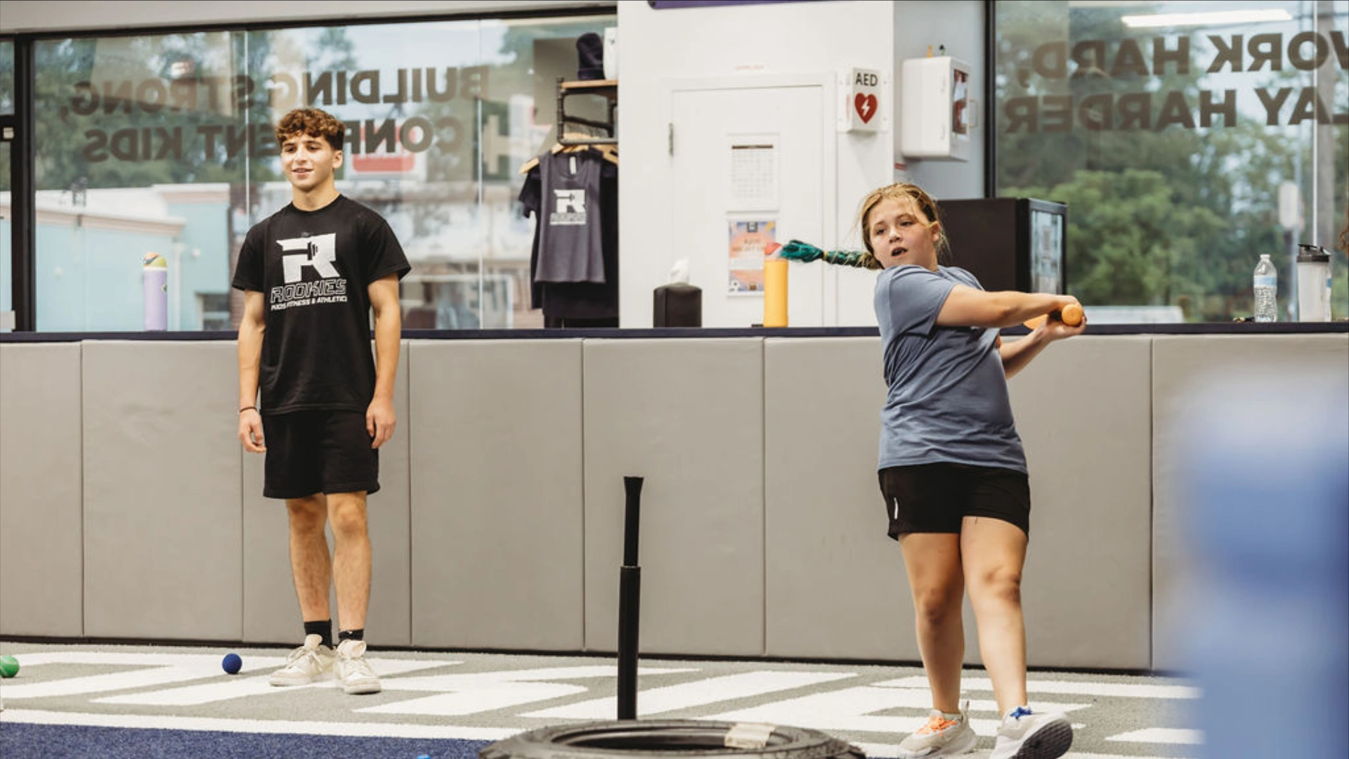 A young girl swings a training bat during a youth sports session at Rookies Kids Fitness & Athletics while a coach watches nearby. The activity takes place on indoor blue turf with batting tees and fitness equipment inside the kids training facility.