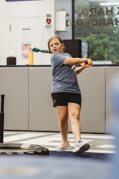 A young girl swings a training bat during a youth sports session at Rookies Kids Fitness & Athletics while a coach watches nearby. The activity takes place on indoor blue turf with batting tees and fitness equipment inside the kids training facility.