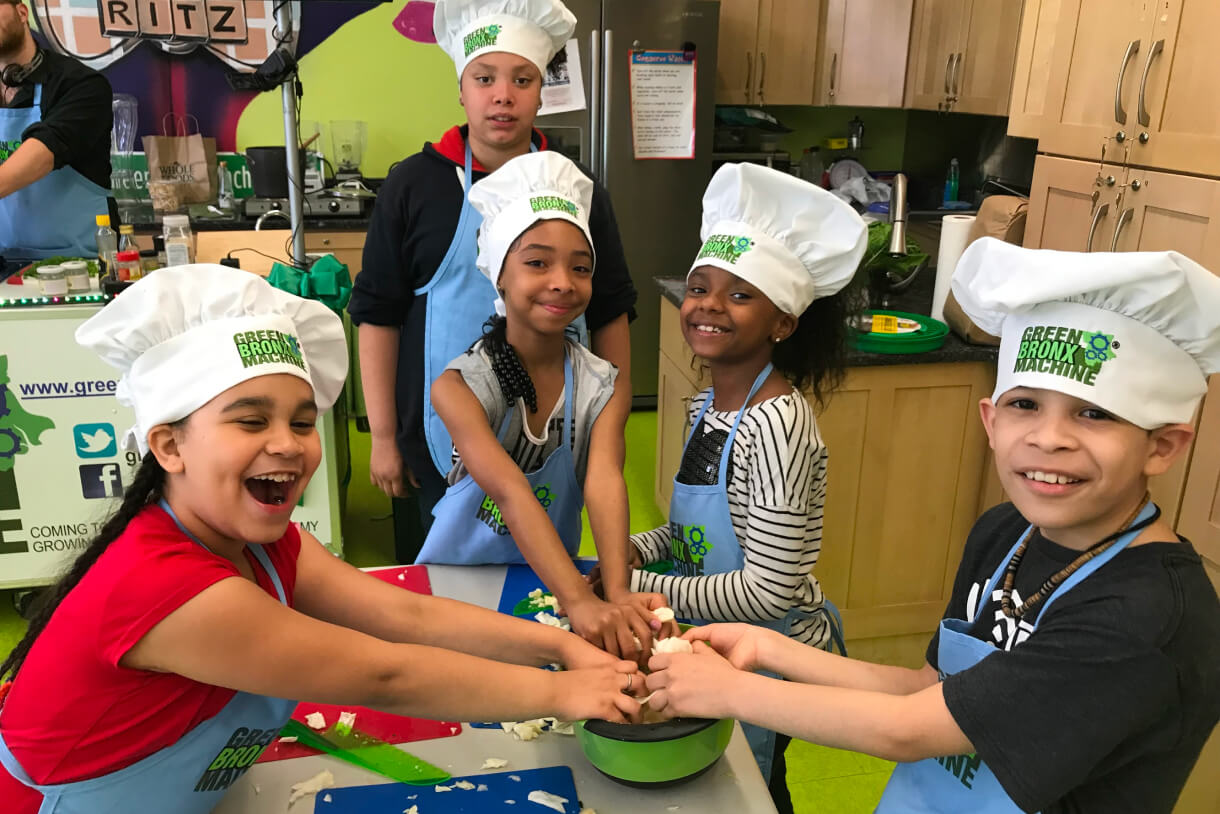 Students in Green Bronx Machine chef hats gathered around a bowl during a classroom cooking lesson