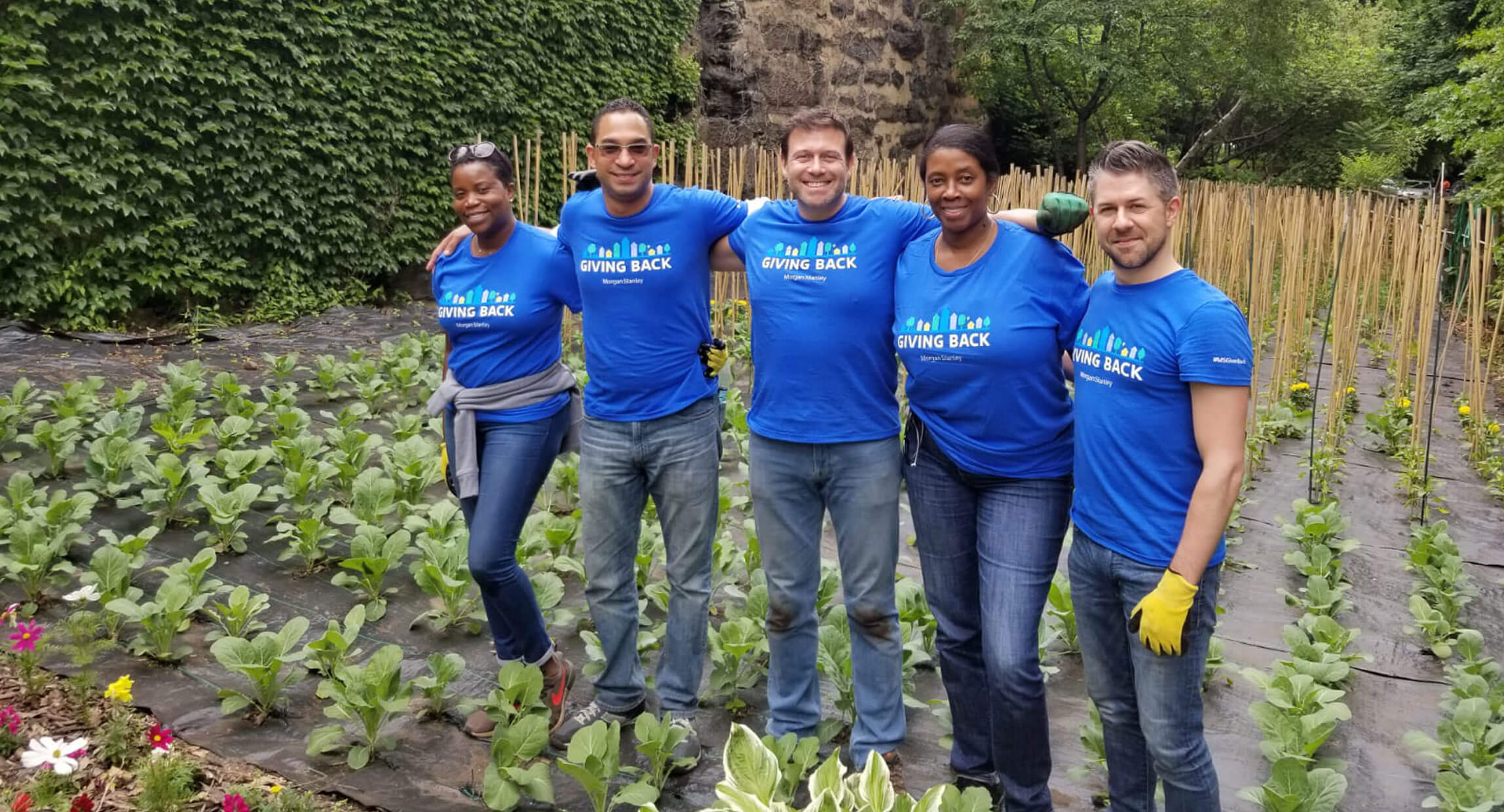 Five volunteers in Giving Back t-shirts standing among garden beds
