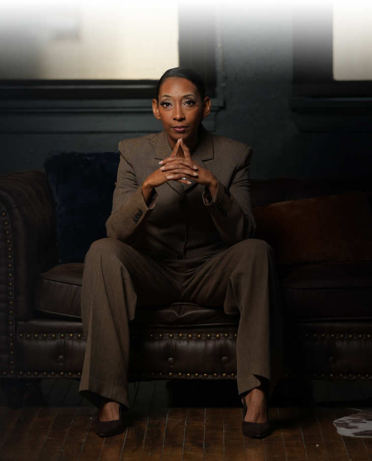 Confident woman in a brown suit sitting on a dark leather couch with hands clasped and elbows resting on knees.