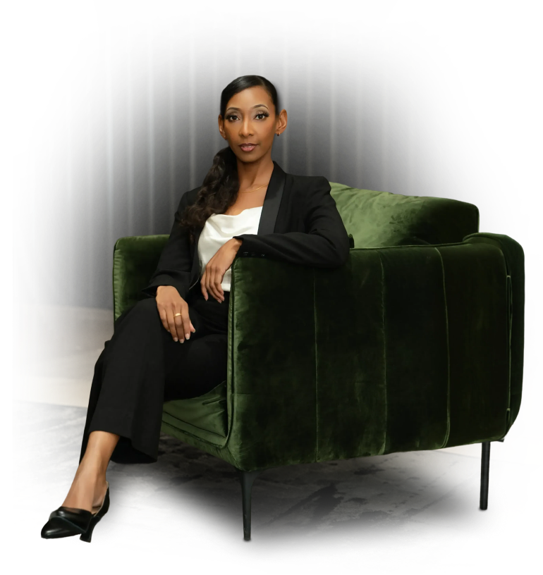 Woman with dark hair in a black blazer and white top sitting confidently on a green velvet armchair.
