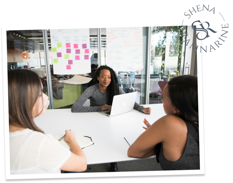 Three women having a discussion around a table in an office, with one using a laptop and others taking notes.