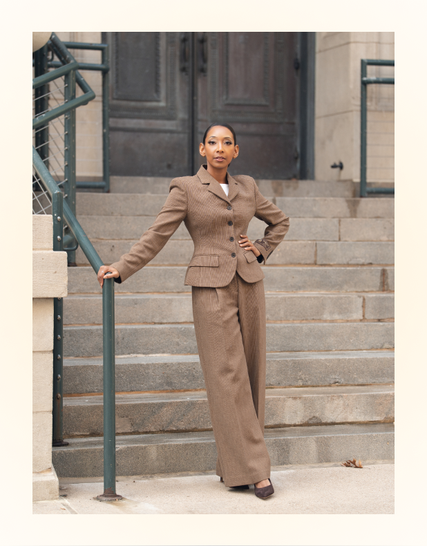 Woman in a brown pantsuit standing on outdoor stone steps, leaning on a green handrail with one hand on her hip.