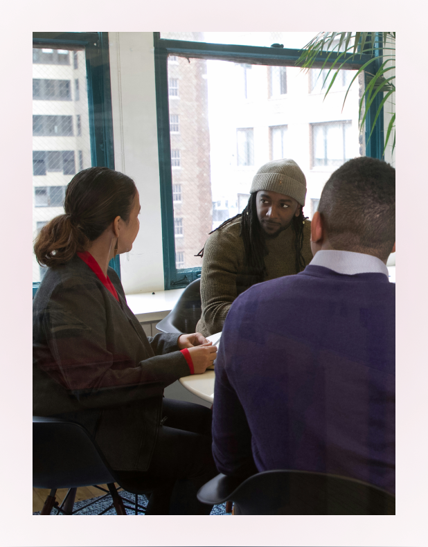 Three people having a conversation around a table in an office with large windows overlooking buildings.