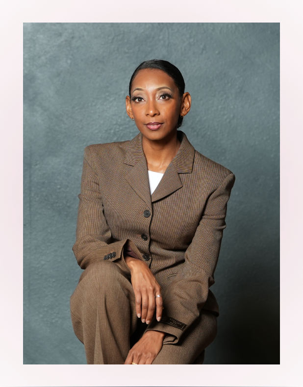 Woman with dark hair tied back, wearing a brown suit, sitting with hands folded against a gray textured background.