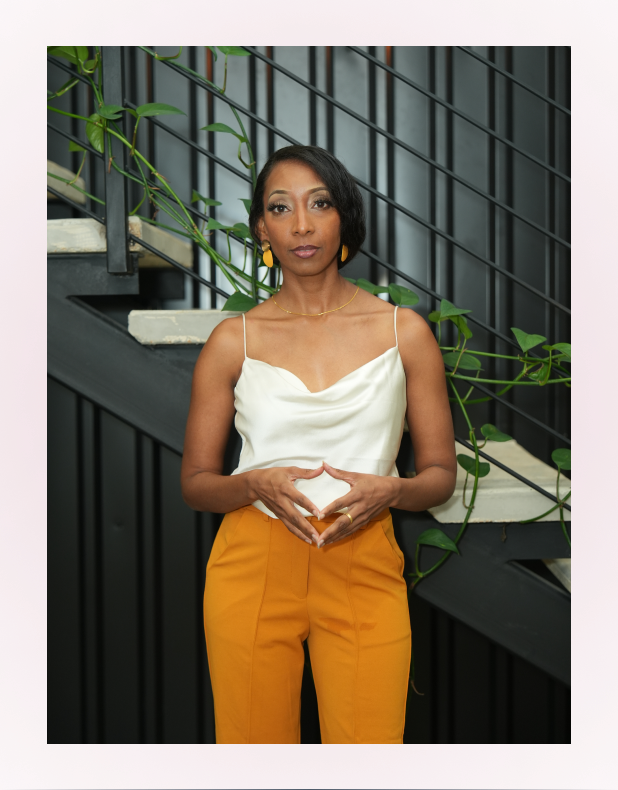 Woman standing in front of a staircase with green plants, wearing a white sleeveless top and mustard yellow pants.