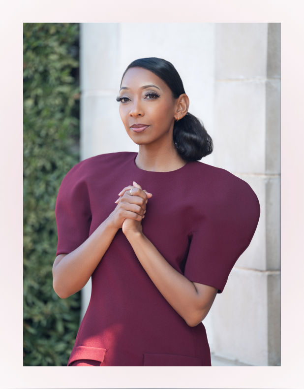 Woman with dark hair pulled back, wearing a burgundy dress with puffed sleeves, standing with hands clasped in front of a stone wall and greenery.