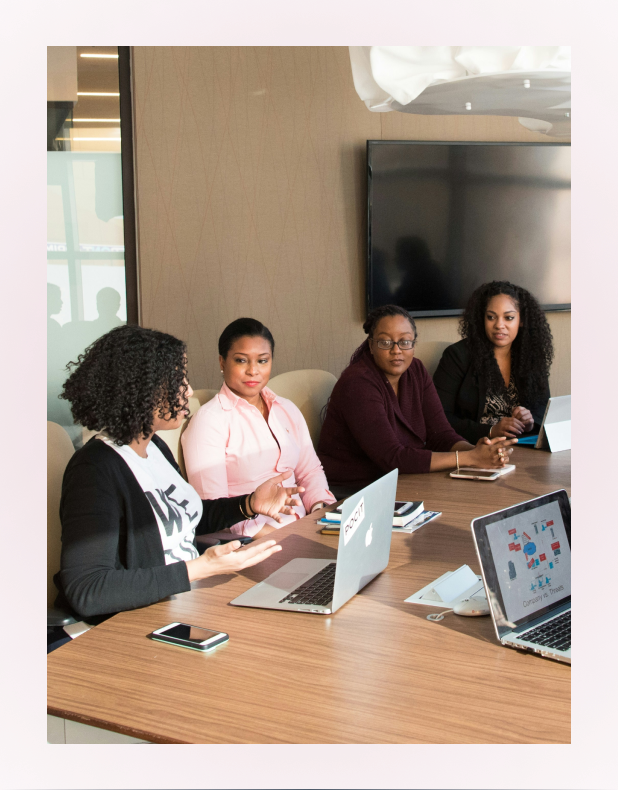 Four women sitting at a conference table engaged in a discussion, with laptops and smartphones in front of them.