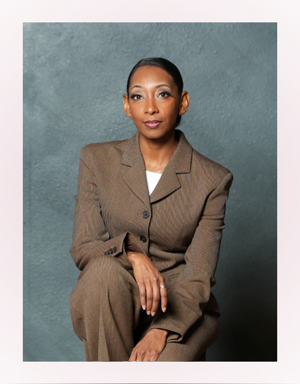 Woman with dark hair tied back, wearing a brown suit, sitting with hands folded against a gray textured background.