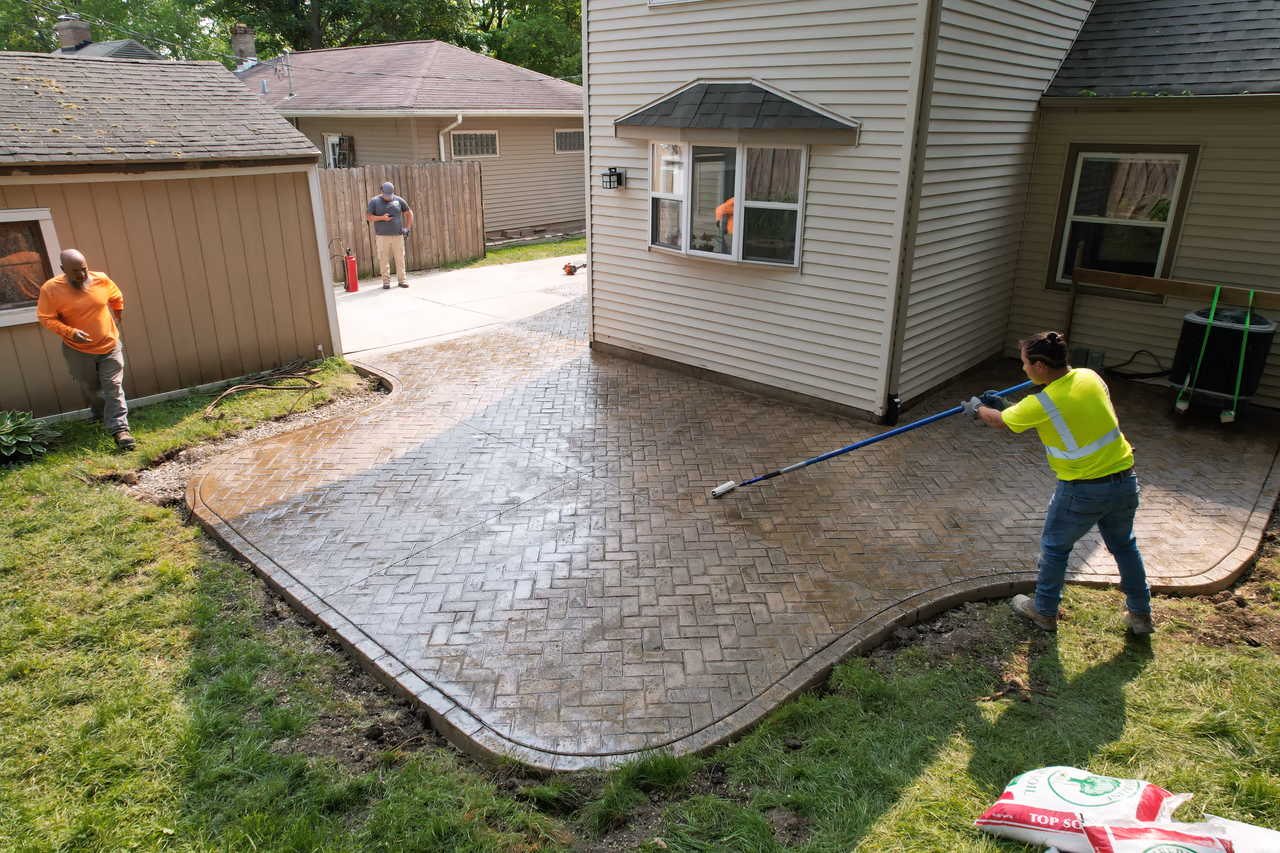 Crew applying sealer to herringbone stamped concrete patio with roller, ground level view