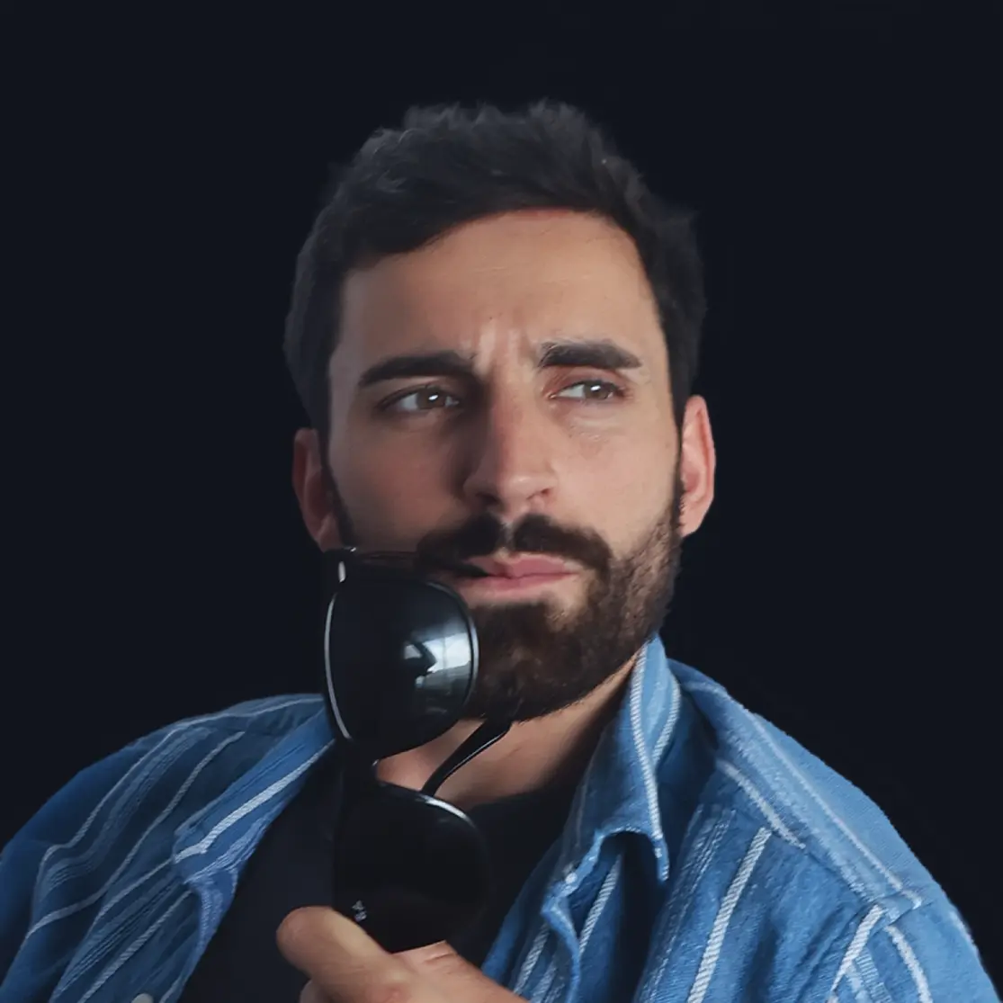 Bearded man with dark hair holding black sunglasses near his chin, wearing a blue striped shirt against a dark background.