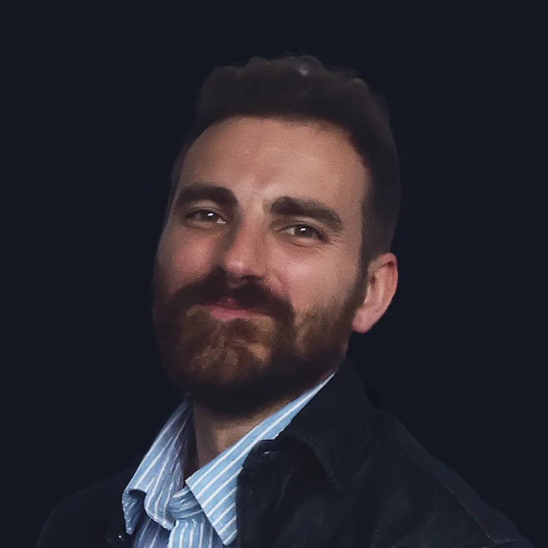 Man with dark hair and beard wearing a blue striped shirt and dark jacket, smiling slightly against a dark background.