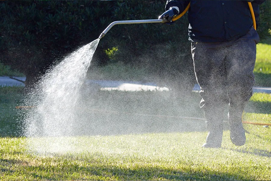 BZ Landscaping technician in dark uniform spraying liquid treatment on a lush green lawn with a handheld wand, creating a fine mist over the grass in a sunny outdoor setting.