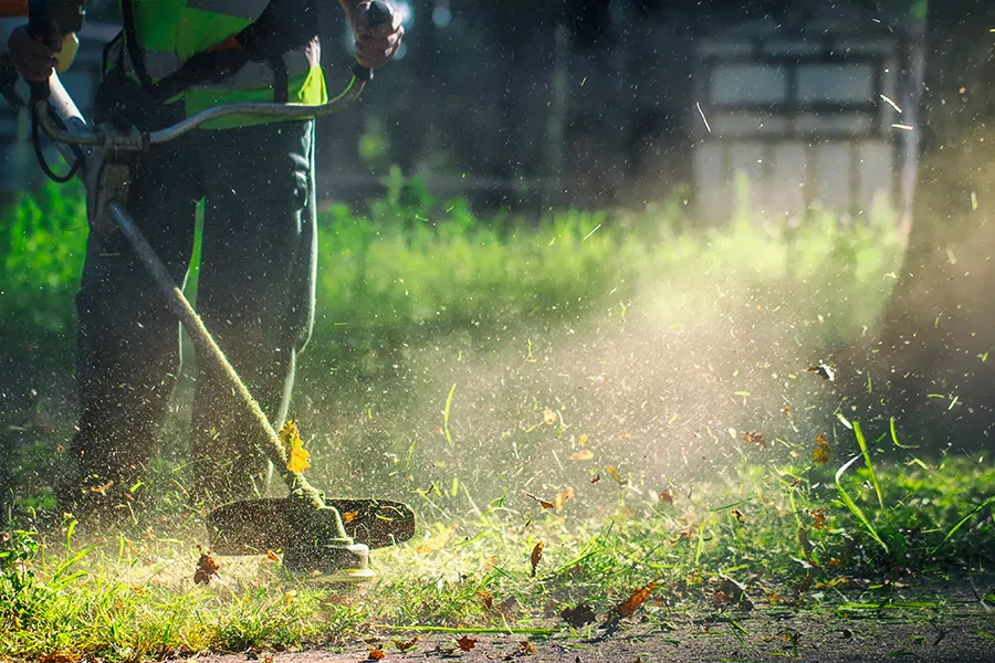 BZ Landscaping worker in a high-visibility safety vest operating a string trimmer to edge a lawn, with grass clippings and leaves flying in the air during outdoor lawn maintenance.