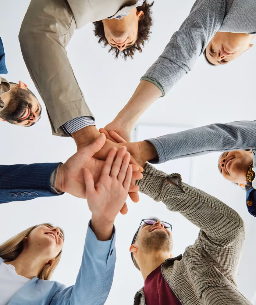Sechs Personen, die im Kreis stehen und sich Hände in der Mitte zusammenschließen als Zeichen für Teamarbeit.