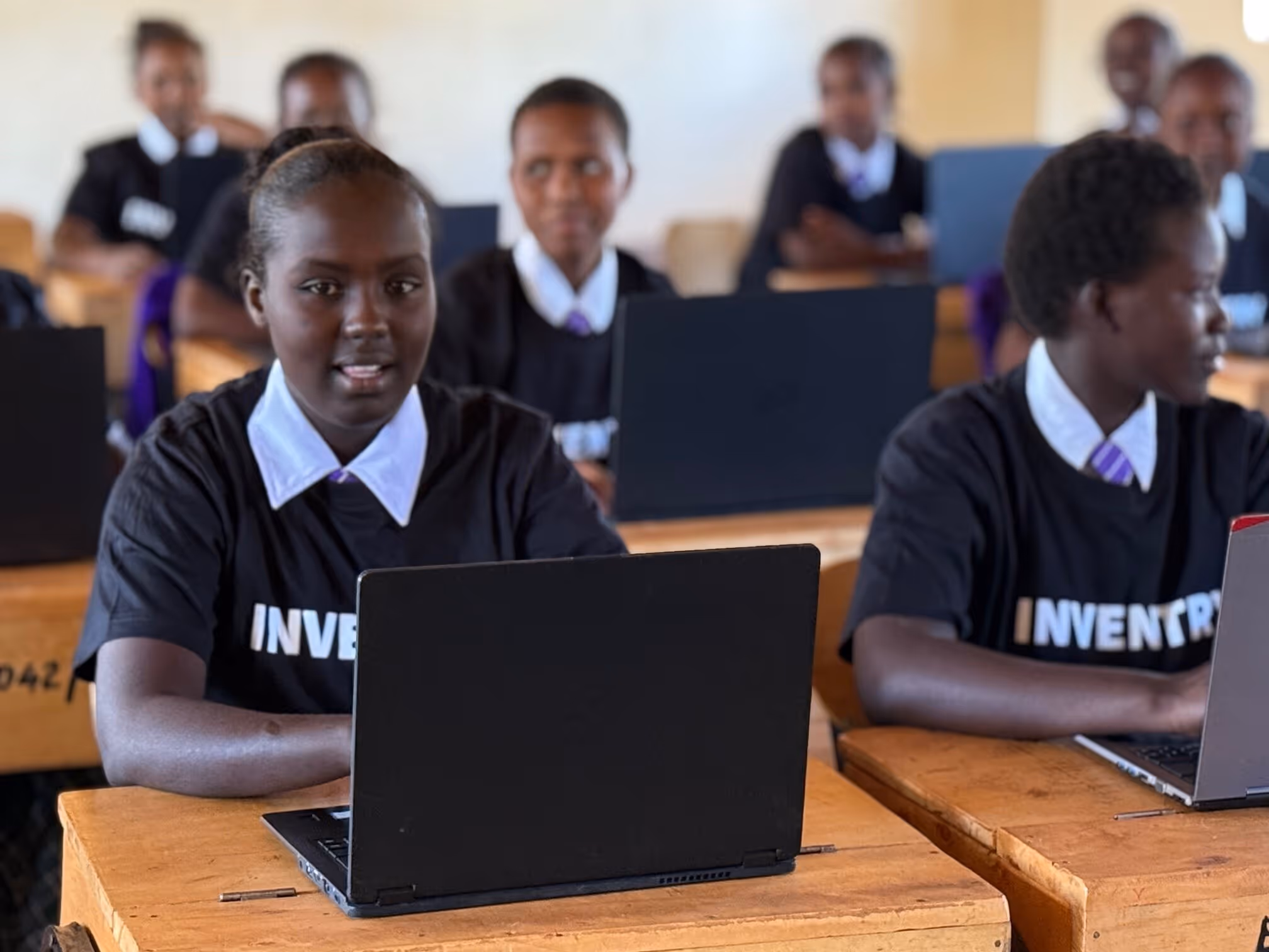 Schülerinnen in Schuluniform sitzen in einem Klassenraum mit Laptops auf Holztischen.