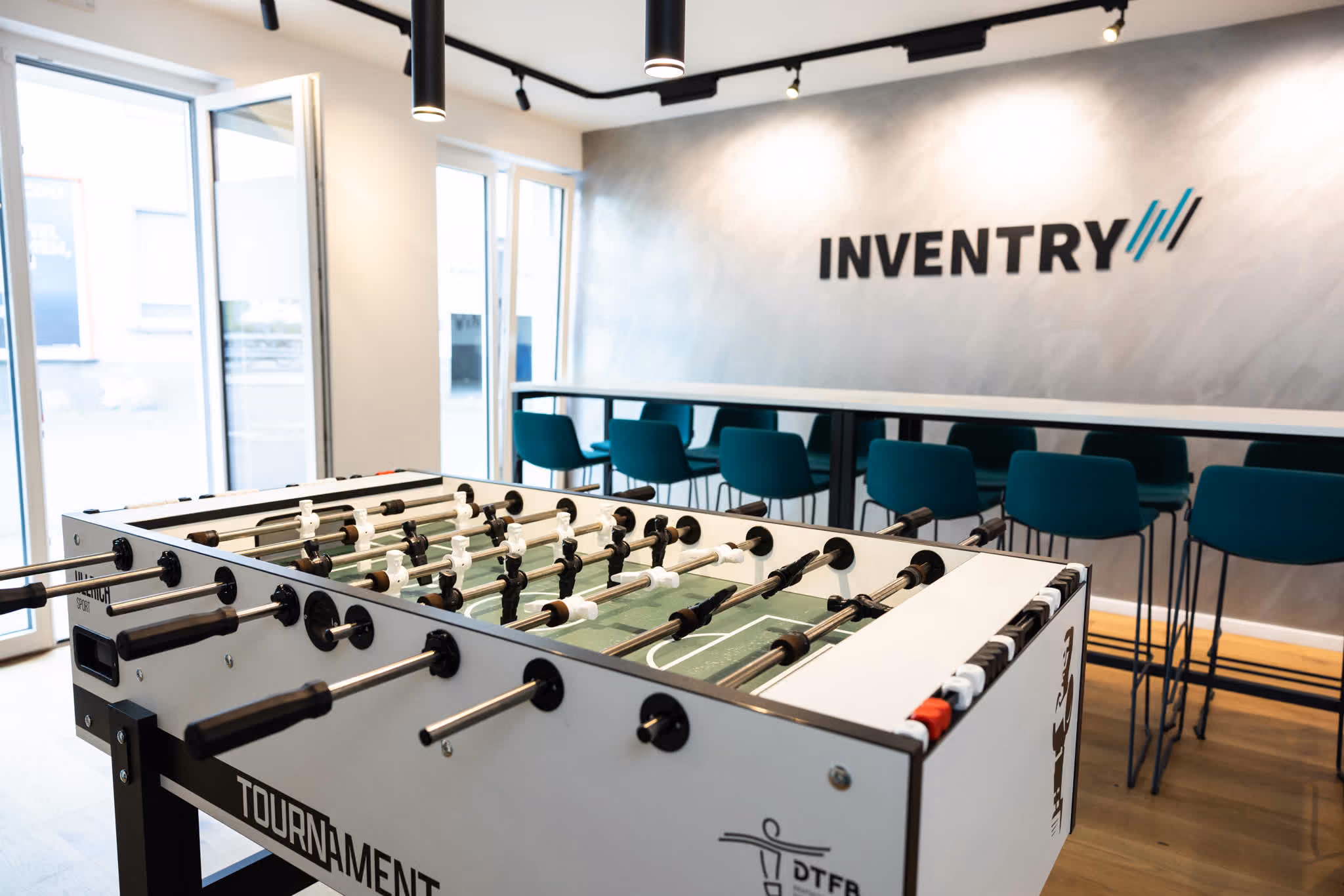 Tischkicker in einem modernen Pausenraum mit blauen Stühlen und INVENTRY Logo an der Wand.