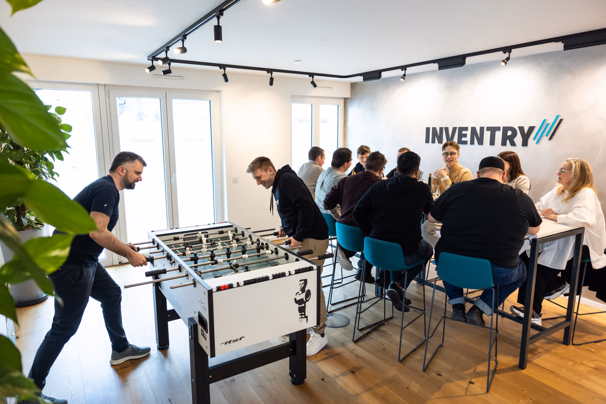 Zwei Männer spielen Tischfußball, mehrere Personen sitzen am Tisch mit INVENTRY-Schriftzug an der Wand.