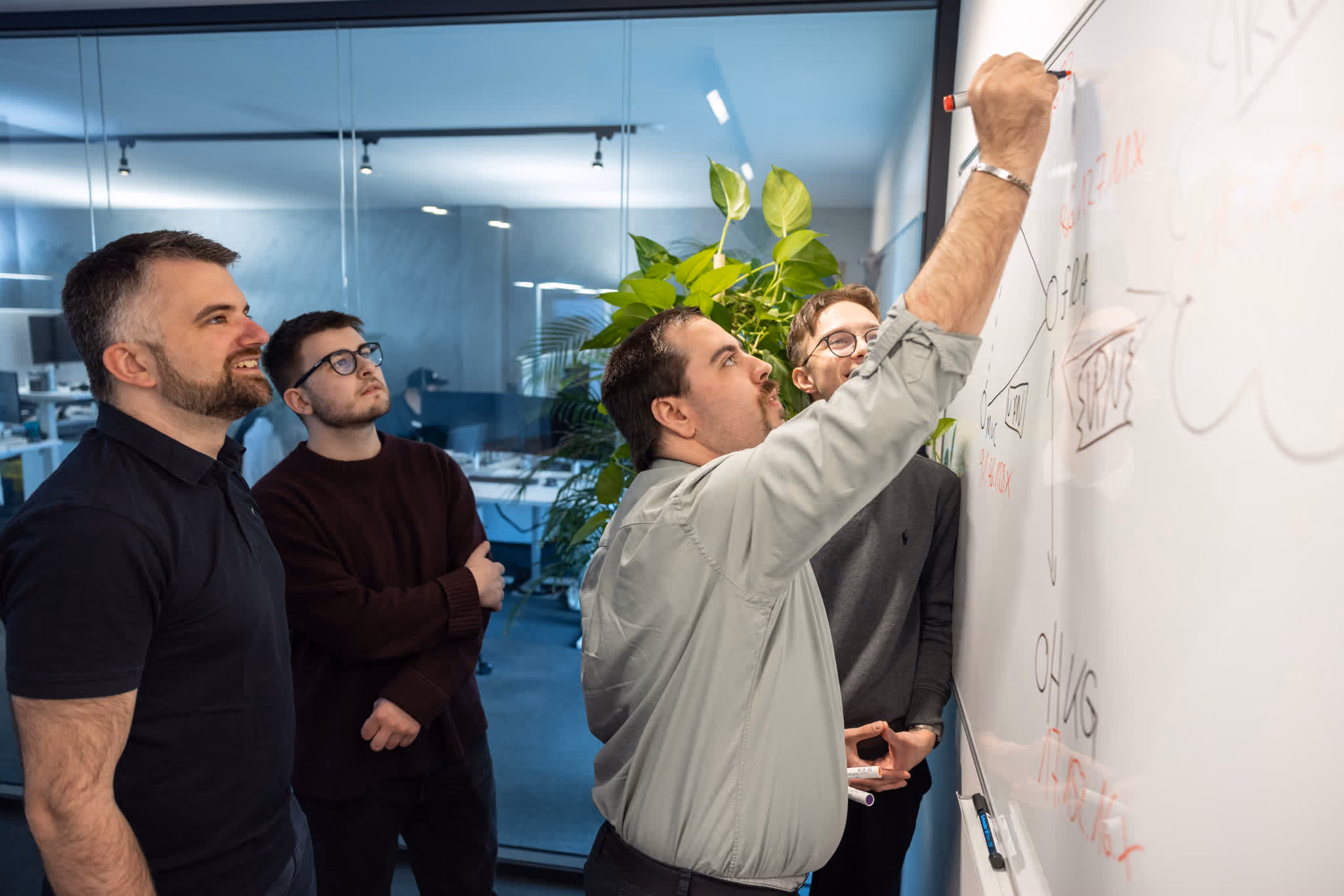 Vier Männer in einem Büro diskutieren vor einem Whiteboard mit Notizen und Diagrammen.
