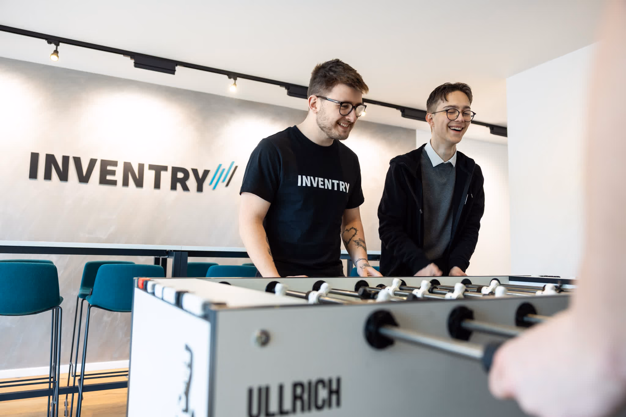 Zwei Männer spielen gemeinsam Kicker in einem modernen Büro mit INVENTRY-Logo an der Wand.