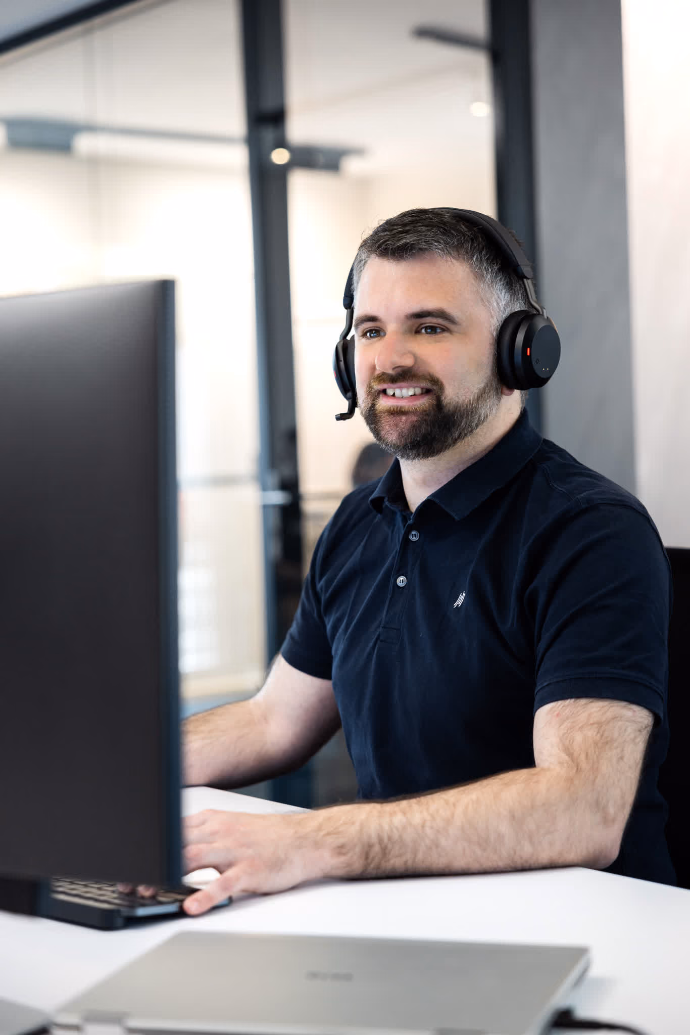 Mann mit Headset lächelt, während er an einem Schreibtisch an einem Computer arbeitet.