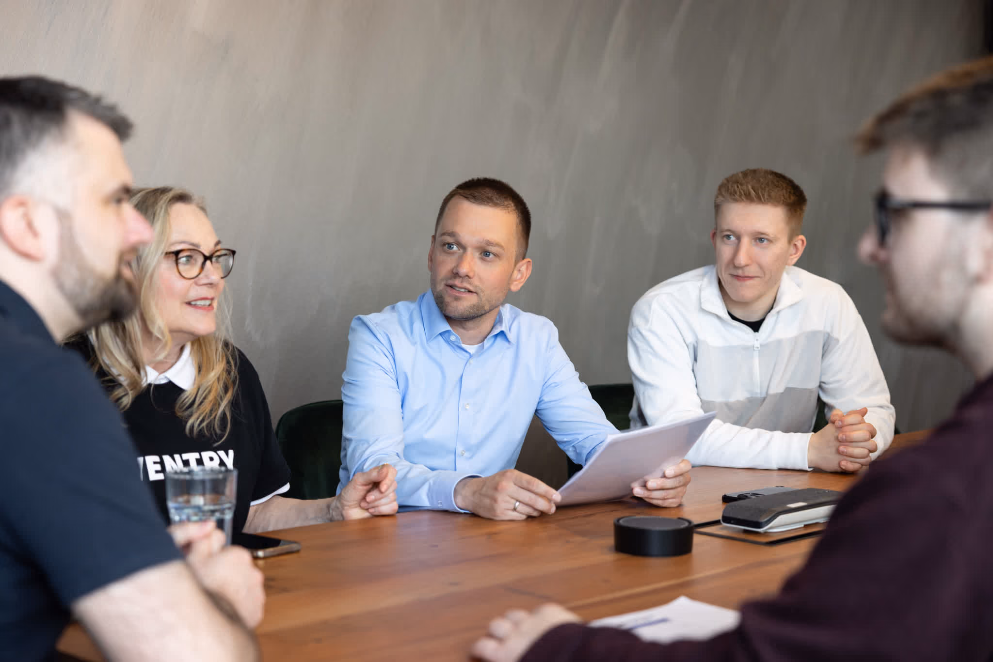 Gruppe von fünf Personen, die bei einem Treffen um einen Holztisch sitzen und sich unterhalten.