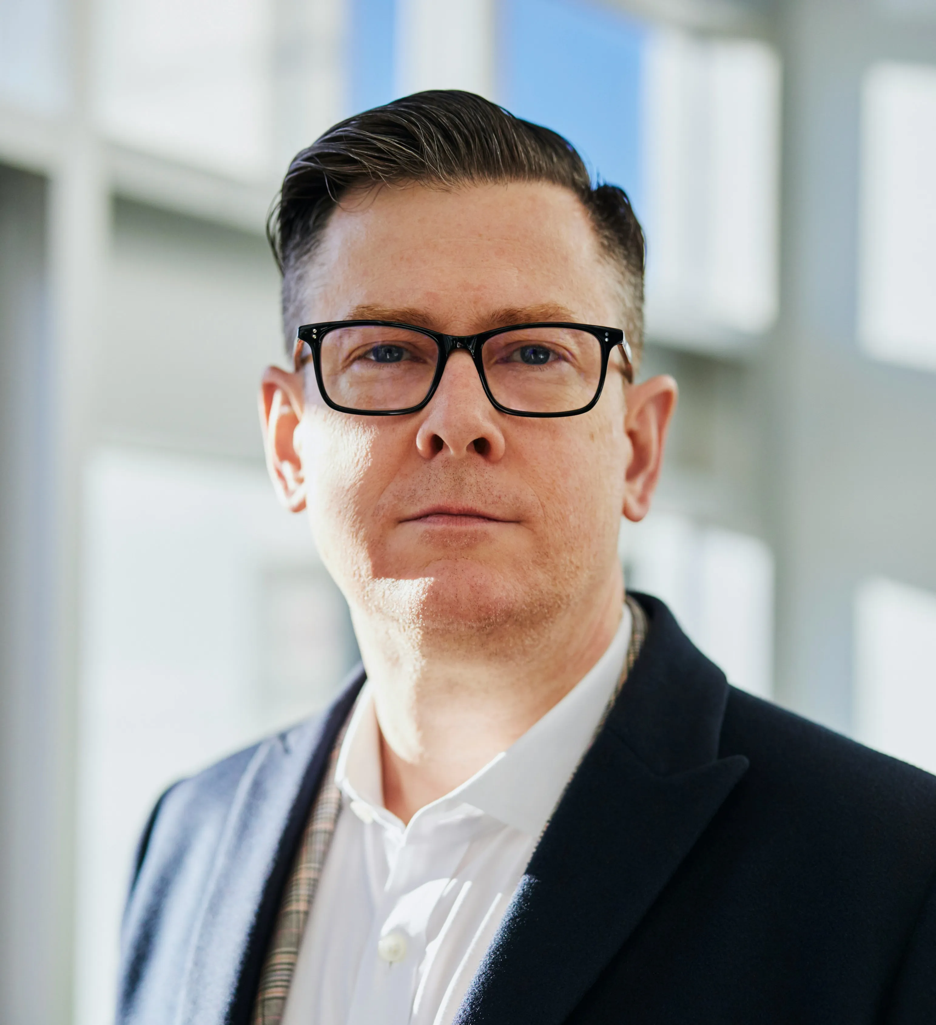 Middle-aged man with glasses wearing a white shirt and dark blazer in soft daylight indoors.