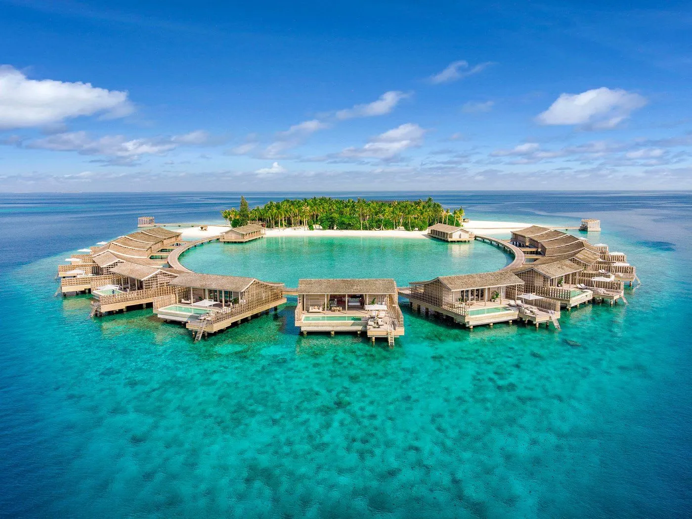 Circular overwater bungalows with pools surrounding a small lush island in clear turquoise ocean.