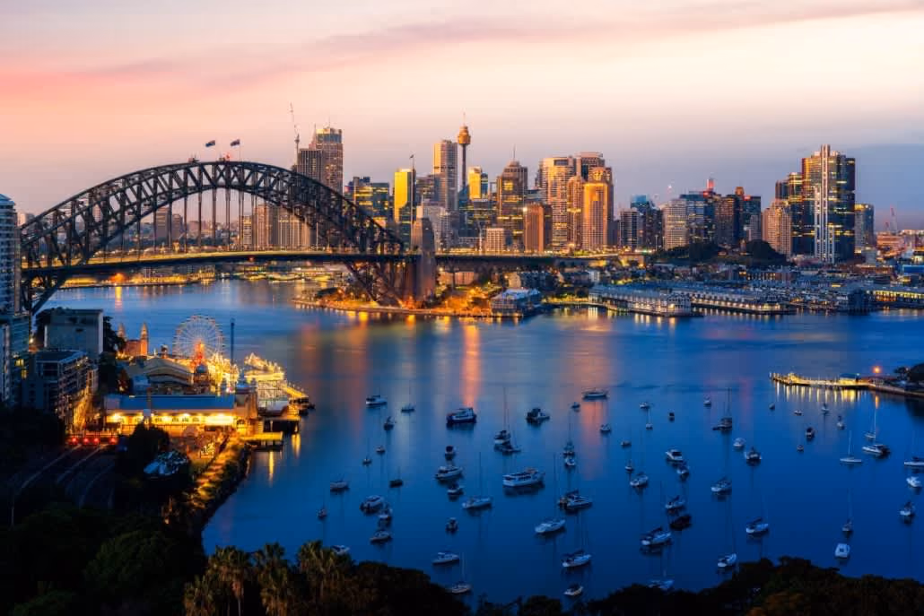 Sydney Harbour Bridge over water with boats and city skyline at sunset.