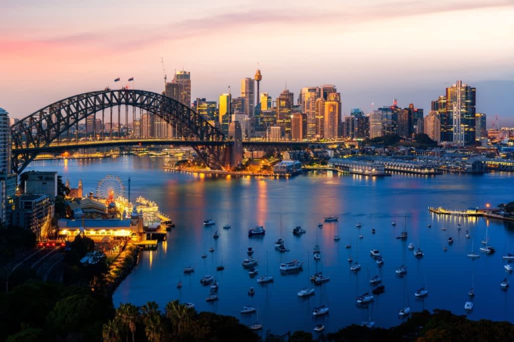 Sydney Harbour Bridge over water with boats and city skyline at sunset.