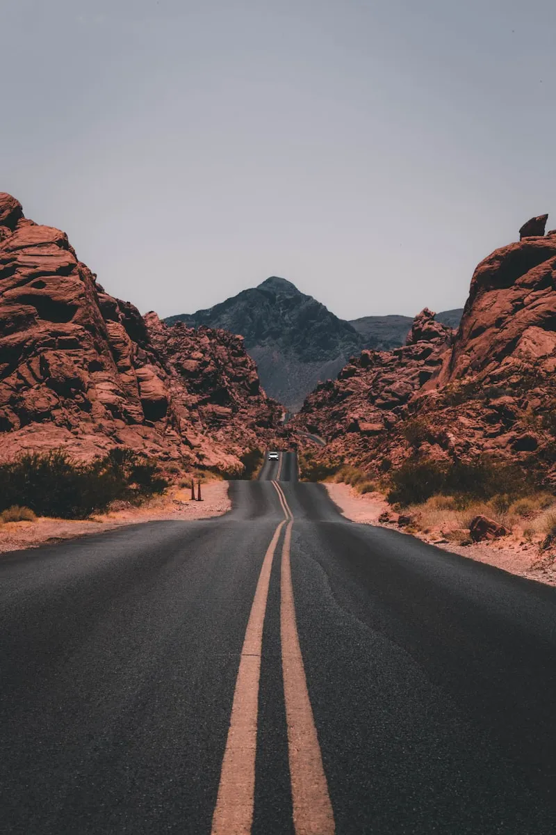 Empty road with double yellow centerlines leading through red rock formations towards a distant mountain.