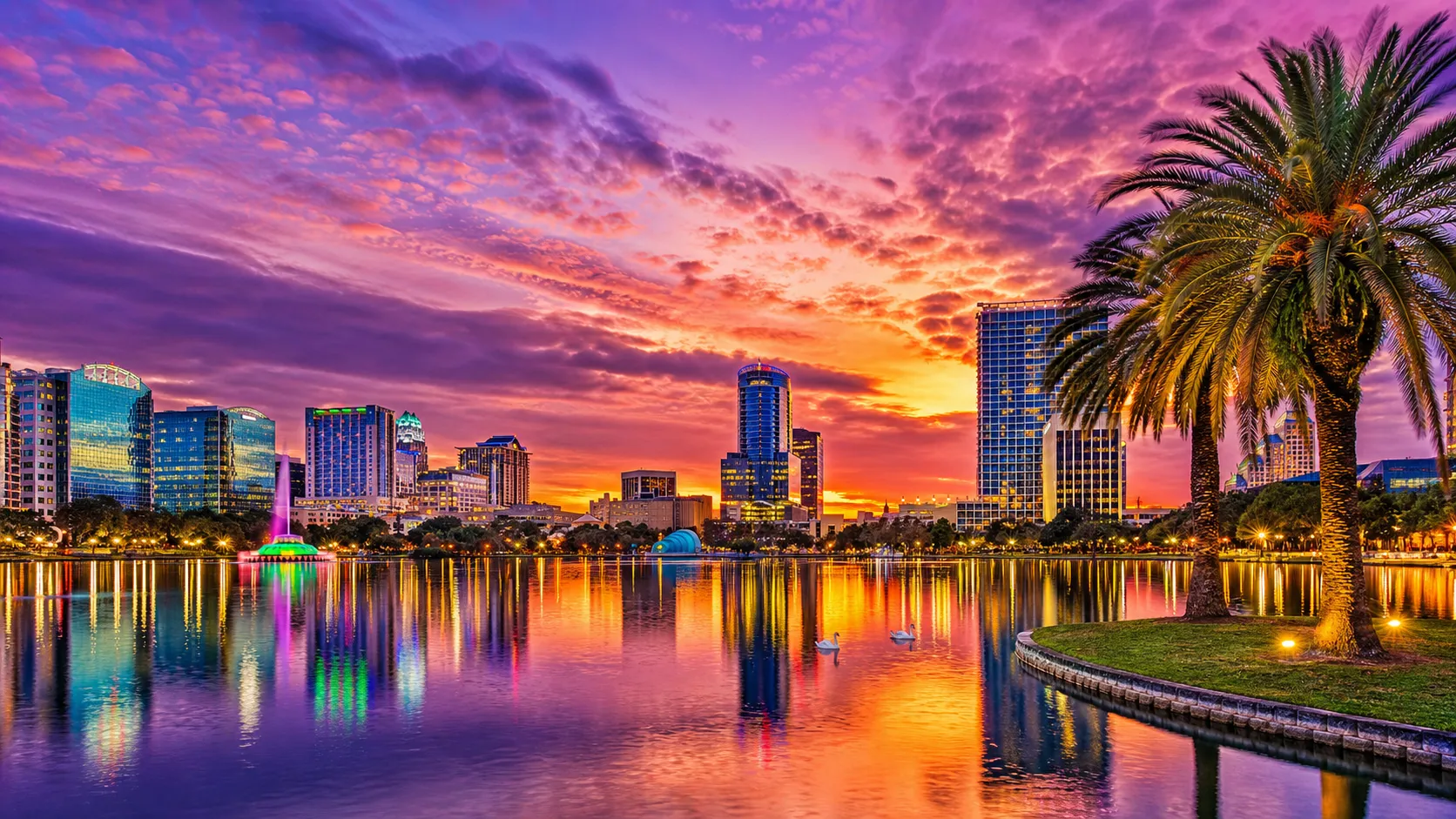 Colorful sunset over city skyline with palm trees and water reflecting vibrant sky colors.