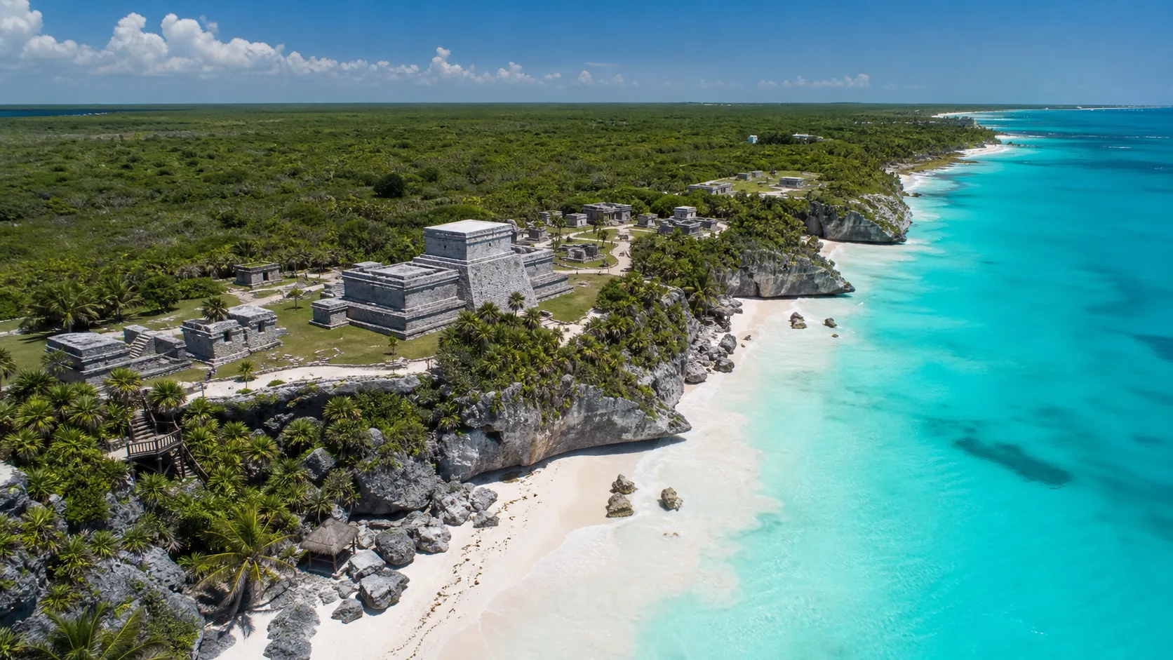 Aerial view of ancient stone ruins on a cliff by a turquoise ocean and white sandy beach.