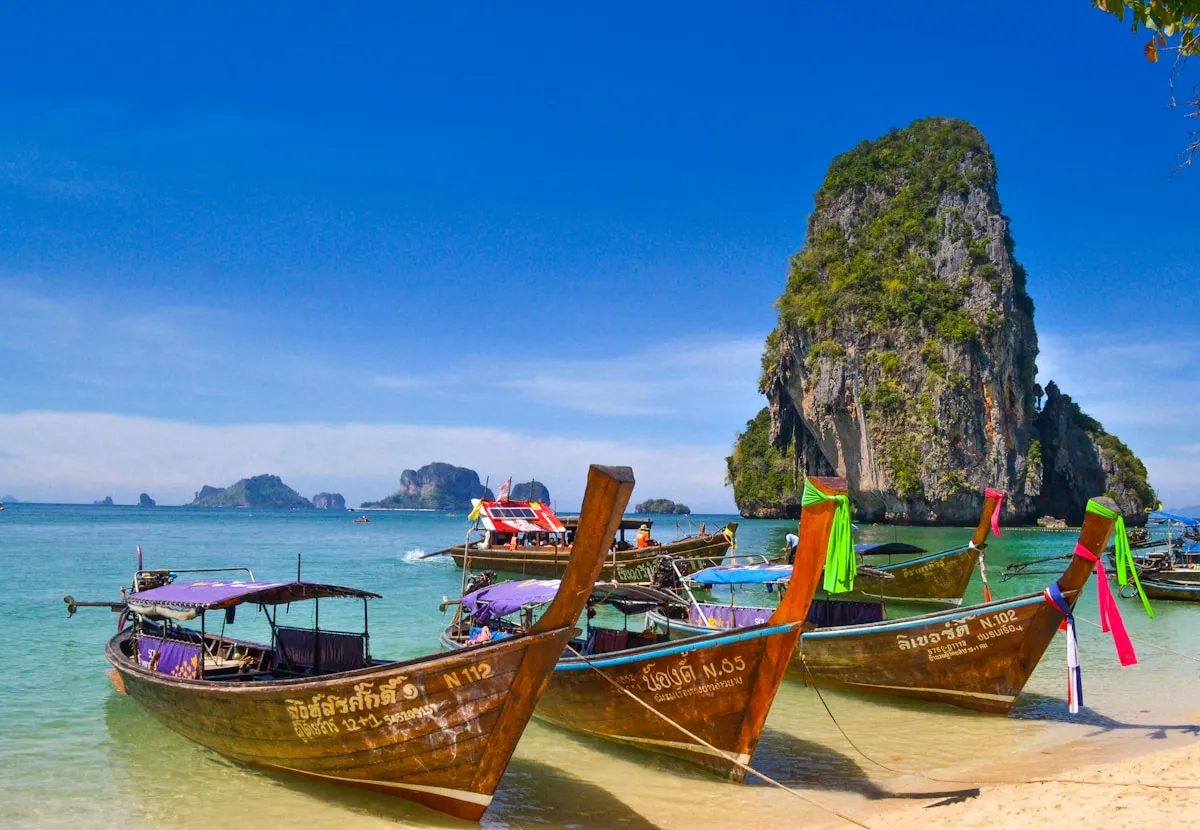 Traditional wooden long-tail boats docked on a tropical beach with clear water and limestone cliffs.