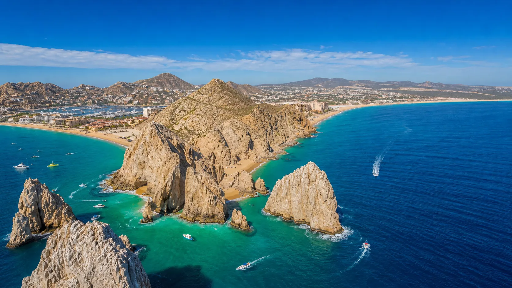 Rock formations in turquoise water near a sandy beach with boats and buildings under a blue sky.