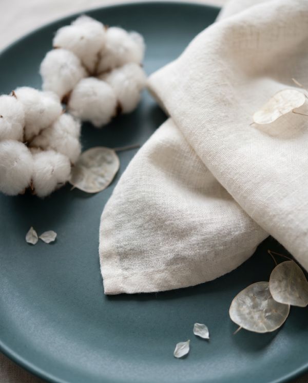 Cotton bolls and natural linen fabric on a matte blue-green plate with translucent dried leaves.