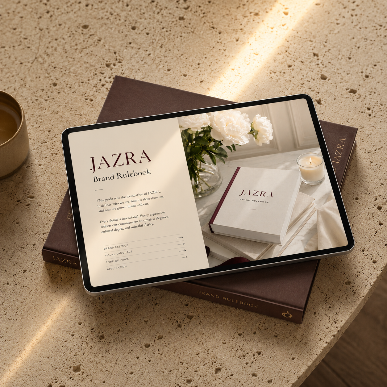 JAZRA Studio Founder Brand Rulebook displayed on a premium workspace with a tablet showing the digital rulebook, styled with white flowers, a candle, and warm natural light.