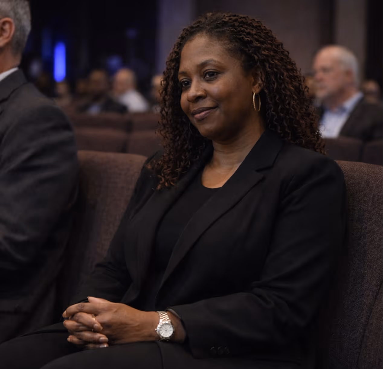 Woman with curly hair wearing a black blazer and watch, seated with hands clasped in a conference.
