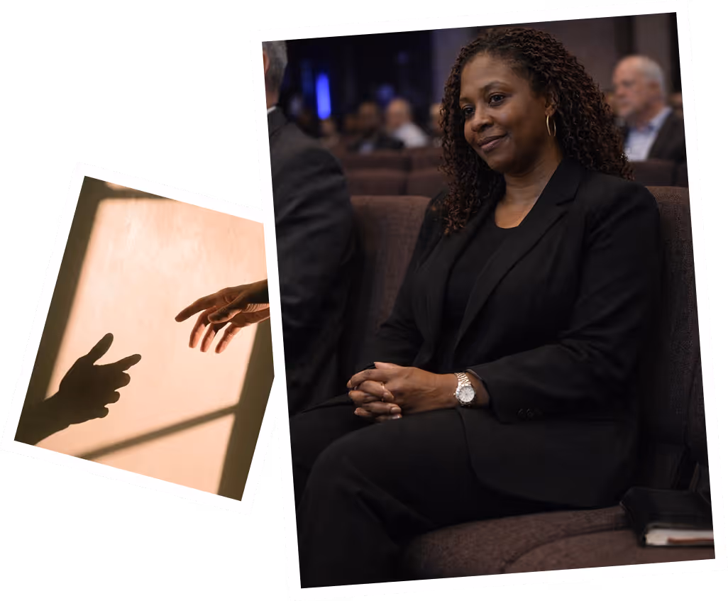 A woman with curly hair in a black suit sitting on a couch in an auditorium with folded hands, beside an image of two hands reaching towards each other in soft light.