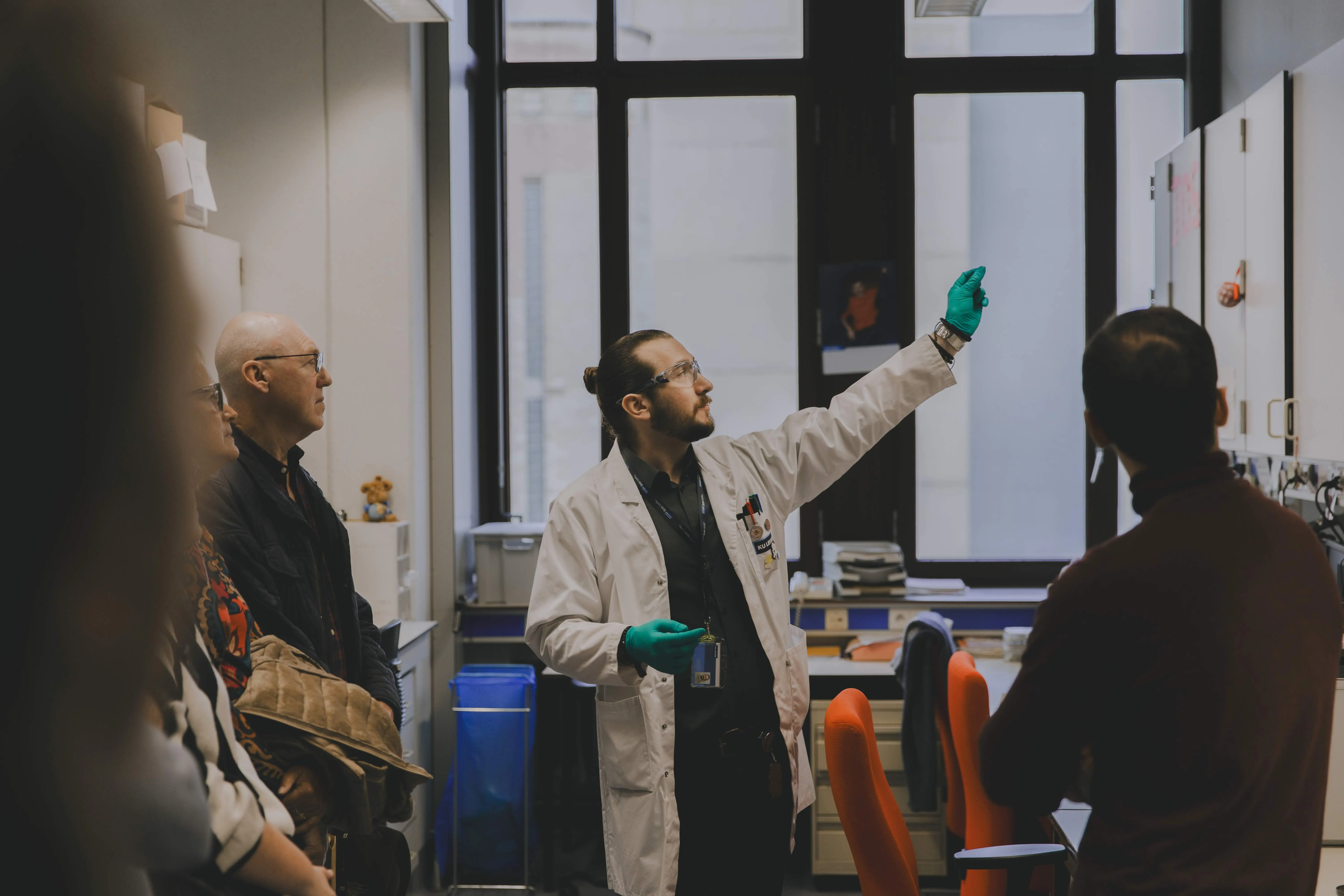 Een wetenschapper in een witte laboratoriumjas en groene handschoenen wijst naar iets boven in een laboratoriumruimte, terwijl meerdere mensen aandachtig naar hem kijken.