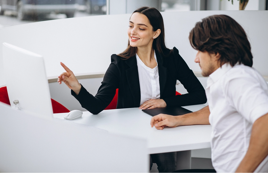 Woman in business attire pointing at a computer screen while explaining something to a man seated beside her at a white desk.