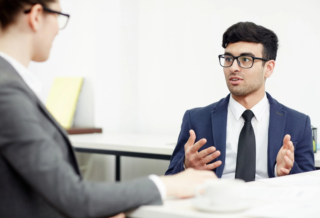 Man in navy blazer and glasses speaking and gesturing with hands during a business meeting with a woman in a gray suit.