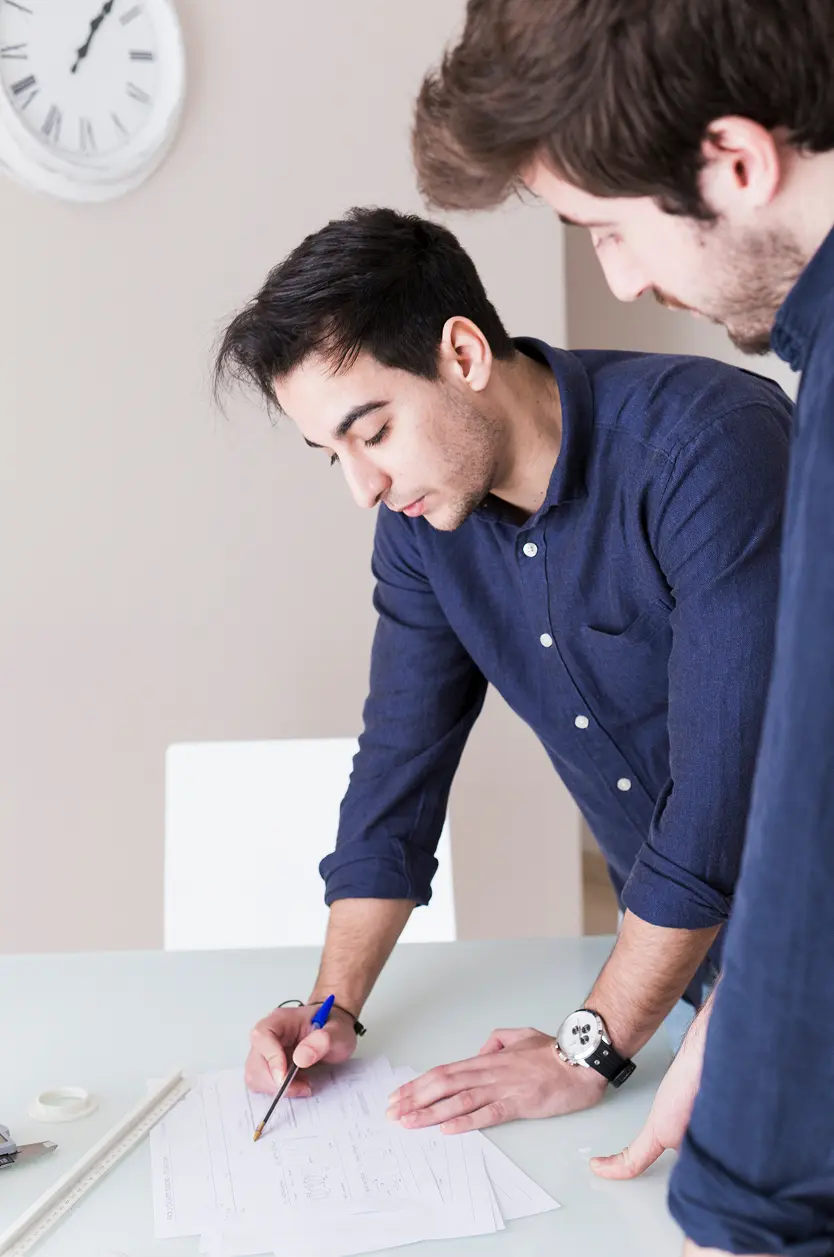 Two men in dark blue shirts collaborate over documents on a table in a bright room with a white clock on the wall.