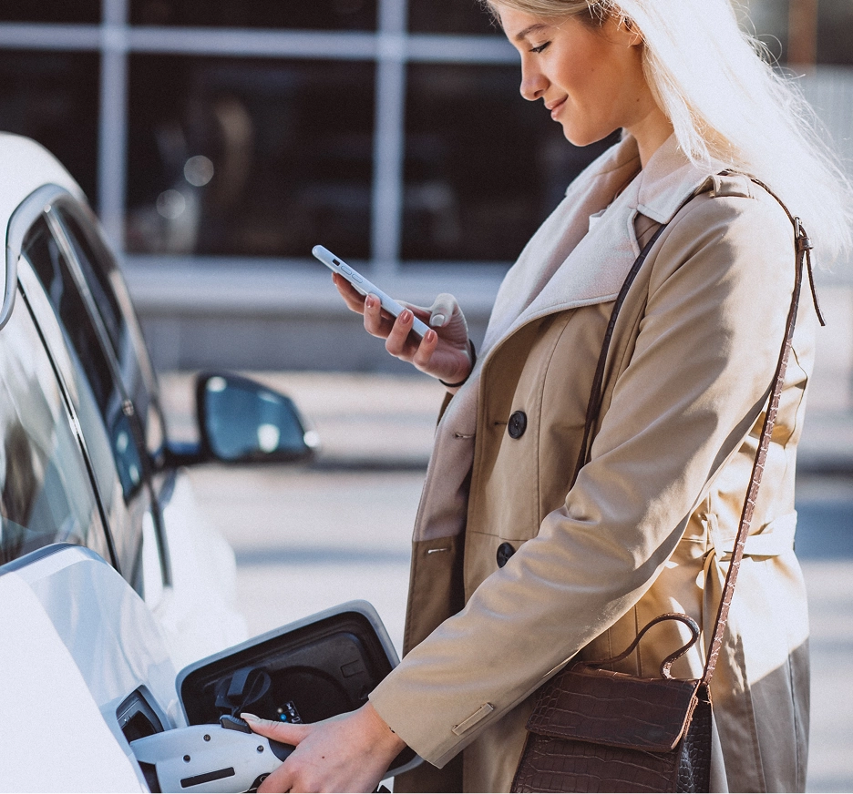 Woman in beige coat using a smartphone while plugging an electric vehicle into a charger.