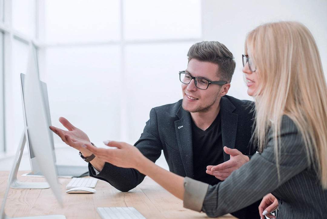 Two coworkers with glasses discussing and pointing toward a computer screen in a bright office.