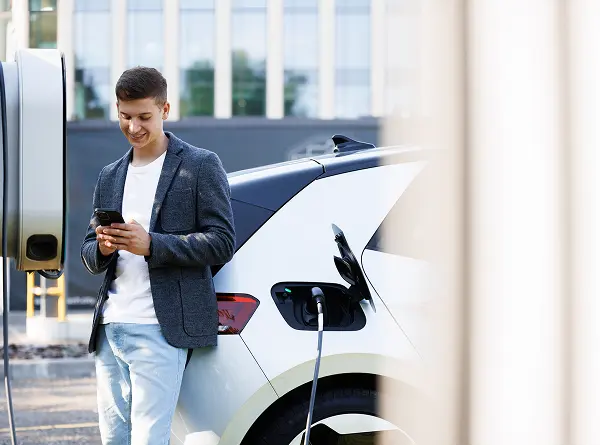 Young man standing by an electric car plugged into a charging station, looking at his phone and smiling.